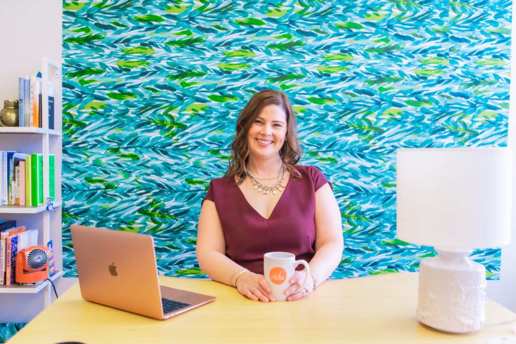 A woman sitting at a desk in front of a bright blue wall with a laptop in front of her