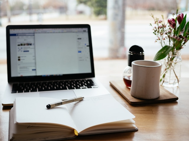 A laptop, notebook, and coffee mug sitting on a bright wooden table