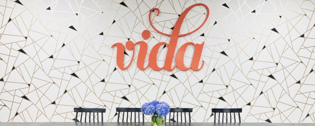 A gold and white patterned wall with the orange VIDA logo on it.