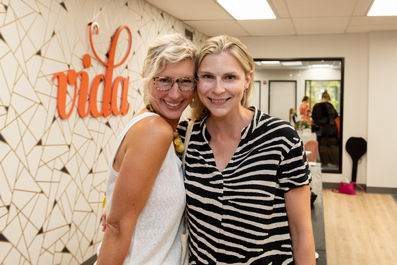 Two blonde women hugging in front of the VIDA logo wall