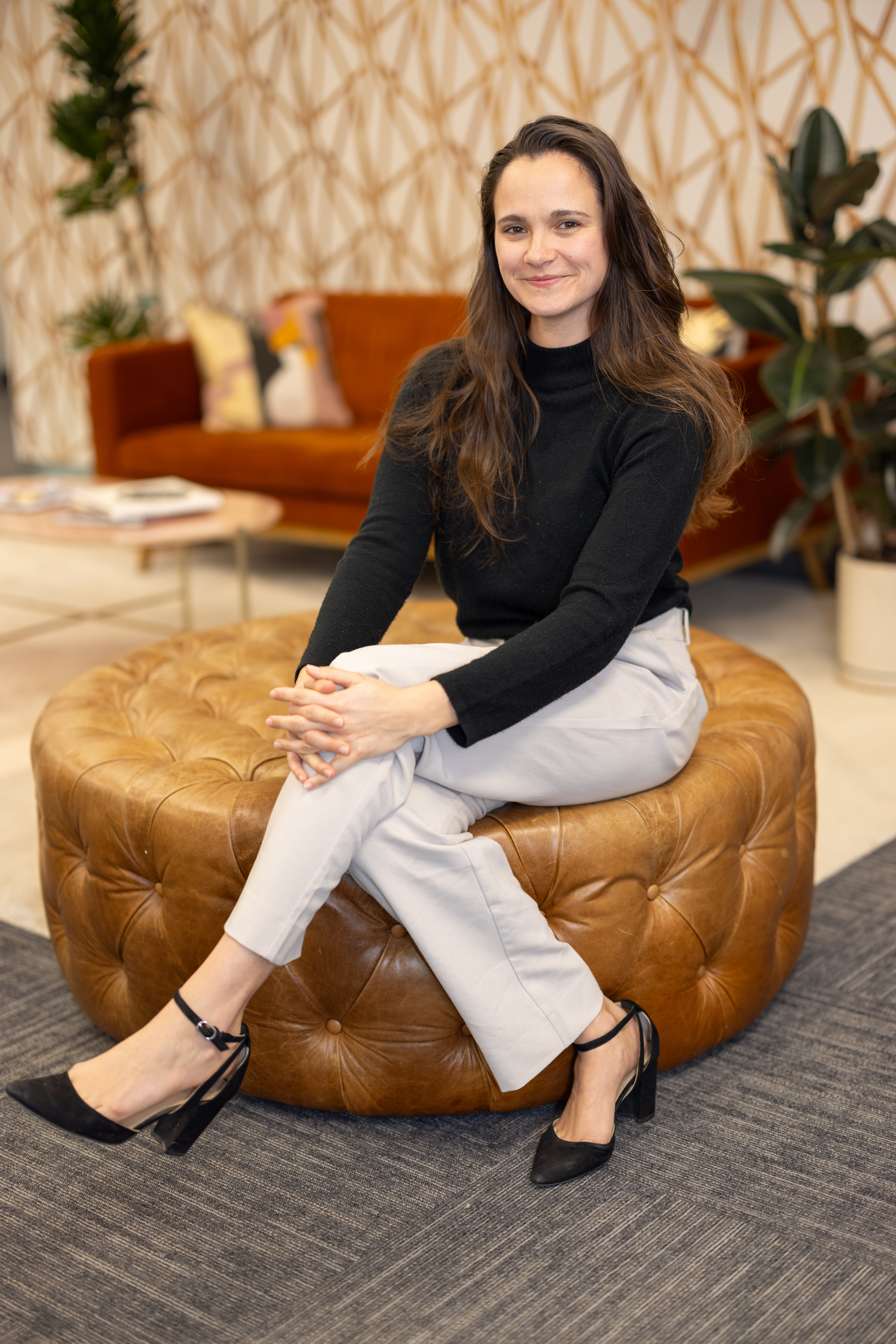 A woman wearing business casual attire sitting on a large gold velvet ottoman