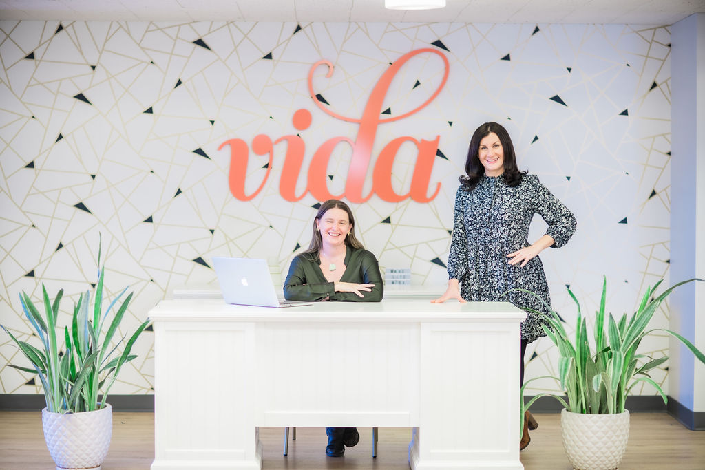 Two woman behind a white desk with the VIDA logo on the wall behind them