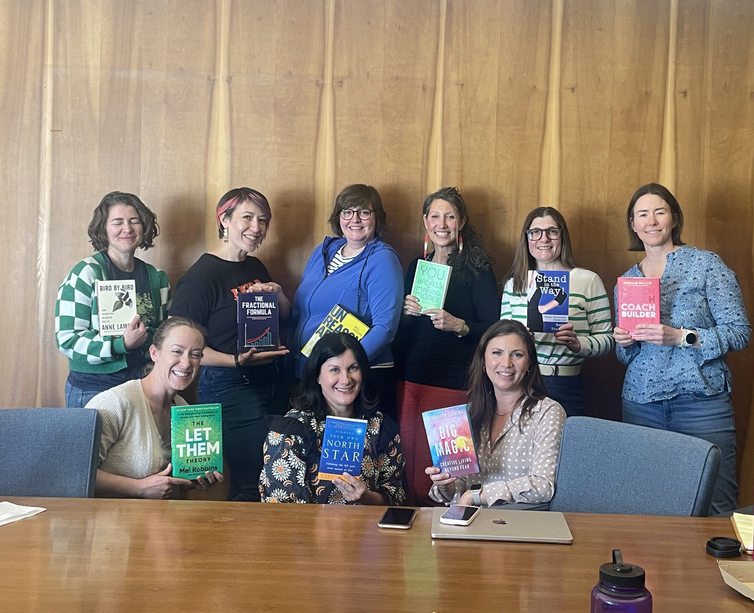 9 women standing behind a conference room table holding books