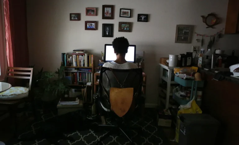 Person viewed from behind working at a dual-monitor desk in a home living room, surrounded by bookshelves, family photos, and a well-lived-in kitchen space in the background.