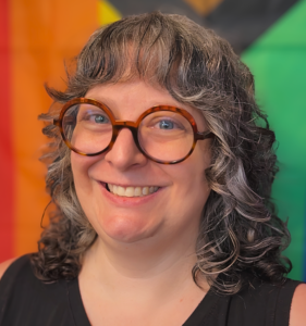 Headshot of Joann, VIDA Virtual Community Manager, smiling in round tortoiseshell glasses against a colorful rainbow-striped backdrop.