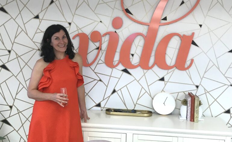 Melanie Marconi, Founder & CEO of VIDA Coworking, smiling in an orange dress in front of the large coral "vida" wall sign at a VIDA location.