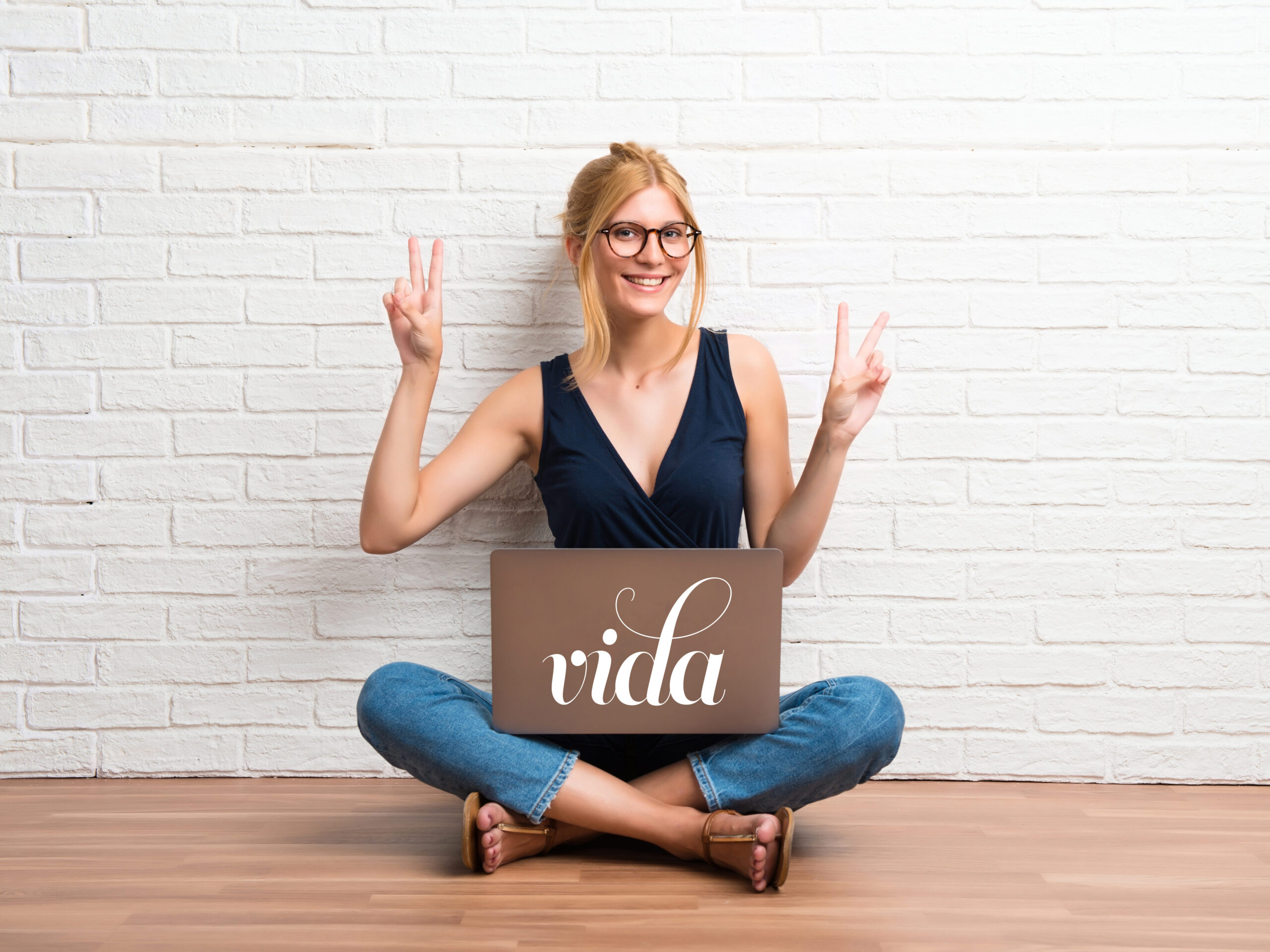 Blonde girl sitting on the floor with her laptop smiling and showing victory sign on white brick wall background